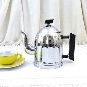May include: A chrome teapot with a black handle and a yellow teacup and saucer on a white lace tablecloth.