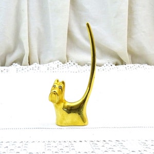 May include: Gold-colored metal ring holder shaped like a dog with rhinestone eyes. The dog is sitting on a white lace surface.