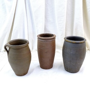 Puede incluir: Tres macetas de cerámica marrón, dos son altas y cilíndricas, una es más corta y tiene un asa. Las macetas están sobre una superficie blanca.