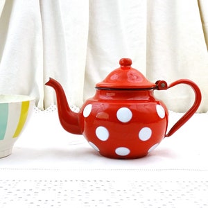 Op de afbeelding: Een rode emaille theepot met witte stippen en een wit deksel. De theepot heeft een gebogen handvat en een tuit. De theepot staat op een wit tafelkleed.