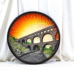 Peut inclure: Une assiette ronde en céramique avec une image 3D d'un aqueduc romain antique. L'aqueduc est peint dans des tons de gris, de brun et de vert, avec un ciel de coucher de soleil en arrière-plan. L'assiette est une pièce décorative de style rustique et vintage.