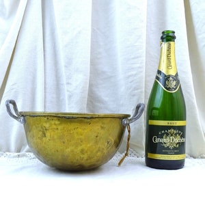 Könnte beinhalten: Eine große, antike Schüssel aus Messing mit zwei Griffen. Die Schüssel steht auf einer weißen Spitzentischdecke. Neben der Schüssel steht eine Flasche Champagner.