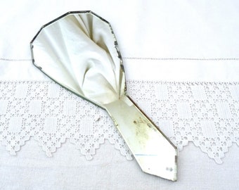 May include: A vintage hand mirror with a faceted, octagonal shape. The mirror is made of glass and has a silver backing. The mirror is lying on a white fabric surface.