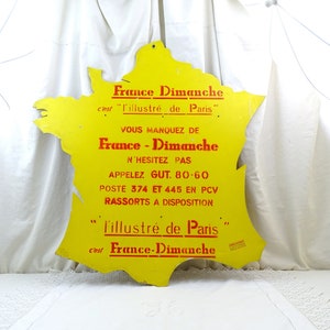 May include: A yellow metal sign shaped like France with red text that reads "France Dimanche" and "l'illustré de Paris". The sign also includes the text "VOUS MANQUEZ DE France - Dimanche N'HESITEZ PAS APPELEZ GUT. 80-60 POSTE 374 ET 445 EN PCV RASSORTS A DISPOSITION" and "l'illustré de Paris cest France-Dimanche".