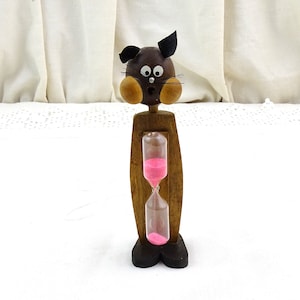 May include: A whimsical wooden hourglass shaped like a surprised cartoon cat. The cat's head is brown with black ears, wooden cheeks, and a surprised expression. The hourglass contains pink sand and is set in a wooden frame.