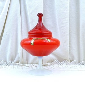 Op de afbeelding: Een rode glazen snoepvaas met een witte voet. De vaas heeft een koepelvormig deksel met een puntige top. De vaas heeft een label met de tekst "Palma Vitrea".