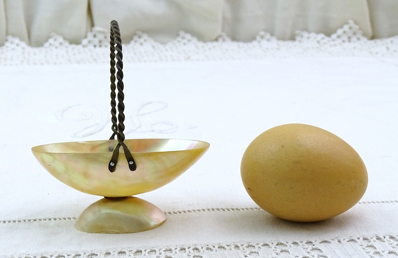 May include: A small, iridescent, mother-of-pearl bowl with a twisted wire handle. The bowl is on a white surface next to a brown egg.