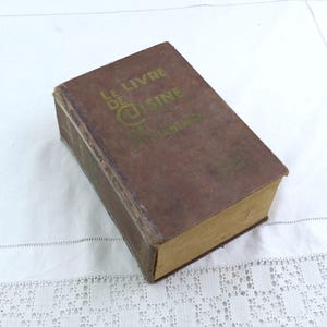 Puede incluir: Un libro de cocina vintage titulado "Le Livre de Cuisine" con letras doradas en una cubierta marrón. El libro tiene un aspecto desgastado, lo que sugiere edad y uso. Las páginas están amarillentas y el libro está sobre una superficie blanca.
