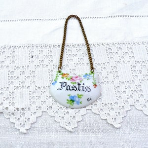 Puede incluir: Etiqueta de botella de porcelana blanca con una cadena dorada. La etiqueta está decorada con flores de colores y la palabra "Pastis" en escritura negra.