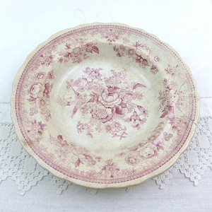 May include: A vintage ceramic plate with a scalloped edge. The plate has a cream-coloured background and a repeating floral pattern in pink hues. The floral design includes roses and other flowers. The plate is round.