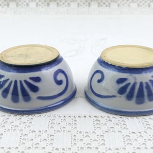 Op de afbeelding: Twee kleine, omgekeerde keramische kommen met een lichtgrijze glazuurlaag en handgeschilderde blauwe bloem- en krulpatronen. Elke kom heeft een ronde, ongeglazuurde beige basis aan de bovenkant en een glad, afgerond buitenoppervlak met blauwe rand.