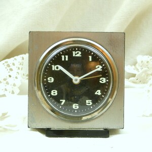 Puede incluir: Un reloj despertador cuadrado plateado con una esfera negra y n&uacute;meros blancos. El reloj tiene una aguja de segundos negra y una aguja de minutos dorada. La esfera del reloj dice "Silvox 2".