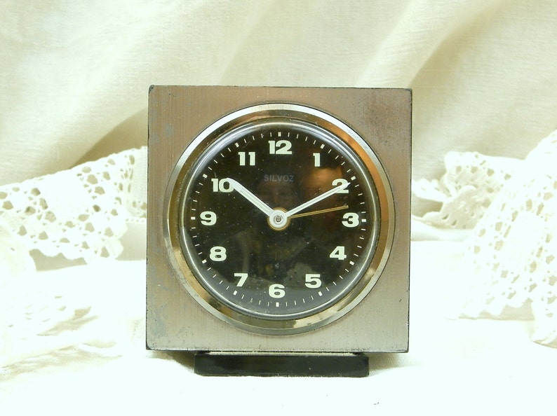 Puede incluir: Un reloj despertador cuadrado plateado con una esfera negra y n&uacute;meros blancos. El reloj tiene una aguja de segundos negra y una aguja de minutos dorada. La esfera del reloj dice "Silvox 2".