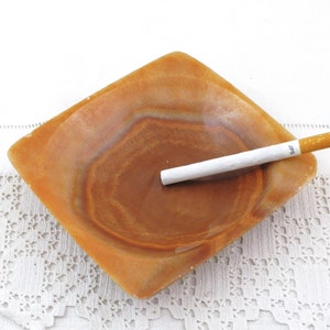 May include: A square, brown, alabaster ashtray with a white cigarette resting inside. The cigarette has the word "JPS" printed on it.