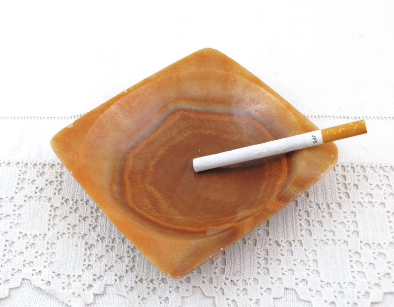 May include: A square, brown, alabaster ashtray with a white cigarette resting inside. The cigarette has the word "JPS" printed on it.