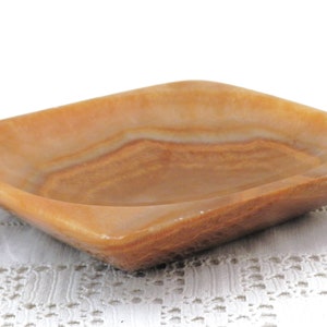 May include: A square, light brown, onyx dish with a slightly raised rim. The dish has a natural, striated pattern.
