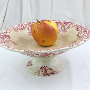 Könnte beinhalten: Eine cremefarbene Keramikschale auf einem Fuß mit einem dekorativen kastanienbraunen Blumenmuster am Rand und am Sockel. Ein einzelner gelb-roter Apfel liegt in der flachen Schale, die einen breiten, ausgestellten Rand und einen stabilen Fuß hat.