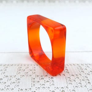 Puede incluir: Un brazalete de forma cuadrada fabricado en resina naranja translúcida y vibrante. El accesorio presenta una superficie lisa y pulida con bordes redondeados y un centro hueco, mostrando un color brillante, uniforme y semitransparente en toda la pieza.