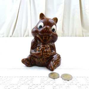 Może przedstawiać: Ceramiczna skarbonka w kształcie brązowej wiewiórki z dużymi białymi oczami i szerokim uśmiechem. Wiewiórka trzyma brązowy żołądź w łapkach i siedzi na białym haftowanym obrusie.