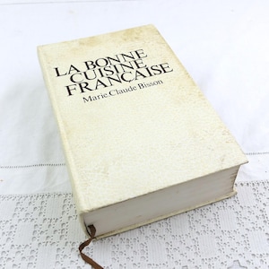 May include: A vintage cookbook titled "La Bonne Cuisine Française" by Marie-Claude Bisson. The book has a cream-colored cover with black text and a brown ribbon bookmark. The book is on a white lace tablecloth.