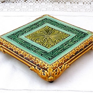 May include: A square ceramic trivet with a green and gold floral design. The trivet has a raised border and four decorative feet.