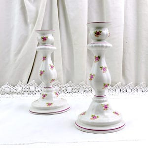 May include: Pair of white ceramic candlesticks with pink rose floral designs. Each candlestick has a flared base, a central bulbous section, and a top cup. The edges are trimmed with a thin, dark pink line.