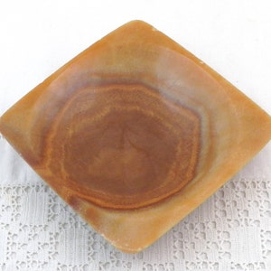 May include: A square, shallow dish made of brown and beige alabaster with a slightly raised rim. The dish has a natural, marbled pattern.