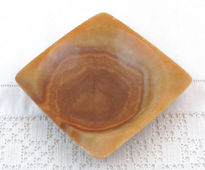 May include: A square, shallow dish made of brown and beige alabaster with a slightly raised rim. The dish has a natural, marbled pattern.