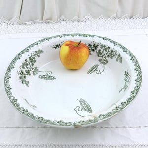 May include: White ceramic serving bowl with a green floral pattern around the rim and interior. A yellow and red apple rests in the center of the bowl. The bowl is on a white tablecloth.