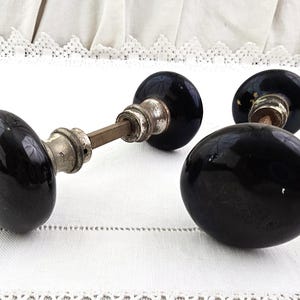 May include: Three antique black door knobs with a glossy finish. The knobs have a rounded shape and are attached to a metal stem with silver-colored hardware. The knobs are displayed on a white surface.