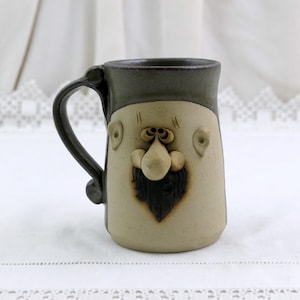 Può includere: Una tazza in ceramica con un design a volto scolpito. La tazza ha una base beige con un bordo, un manico e dettagli del viso grigio scuro. Il volto comprende naso, barba e dettagli degli occhi. La tazza è alta circa 13 cm.