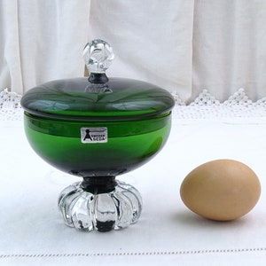 Puede incluir: Un platito de caramelo de vidrio verde con una tapa de vidrio transparente y una base de vidrio transparente. El plato tiene una etiqueta que dice "Sweden Sl&ouml;jd".