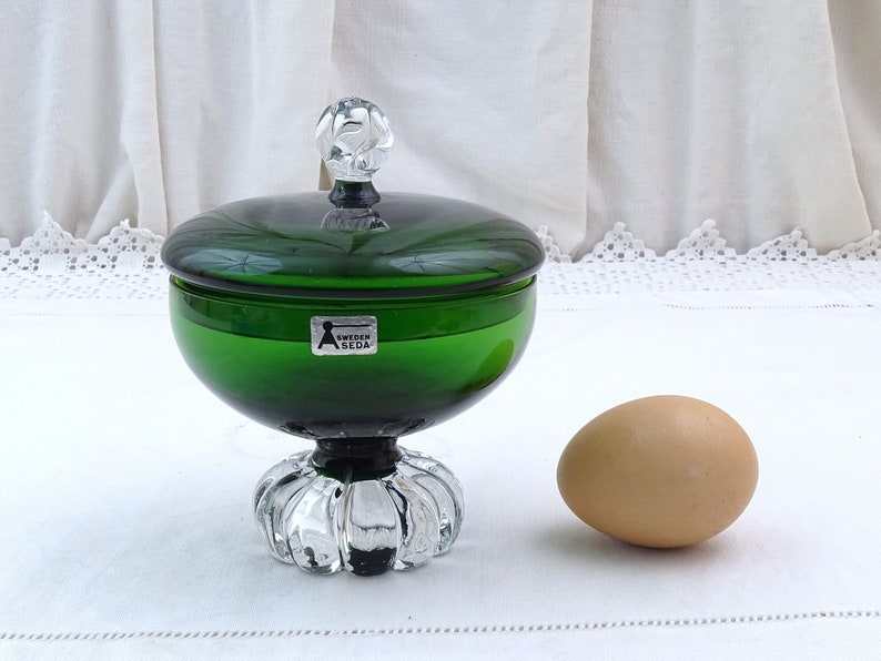 Puede incluir: Un platito de caramelo de vidrio verde con una tapa de vidrio transparente y una base de vidrio transparente. El plato tiene una etiqueta que dice "Sweden Sl&ouml;jd".