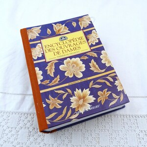May include: A French language book, "Encyclopédie des Ouvrages de Dames" by Thérèse de Dillmont, with a dark blue cover featuring gold floral designs. The spine is brown. The book is a new edition.
