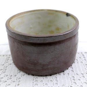 May include: A brown ceramic bowl with a wide rim and a slightly tapered body. The interior is a lighter, off-white color. The bowl sits on a white lace tablecloth. This rustic bowl is suitable for kitchen or home decor.