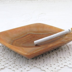 May include: A square, light brown, marble ashtray with a white cigarette resting inside. The ashtray has a slightly raised edge and a smooth, polished surface.