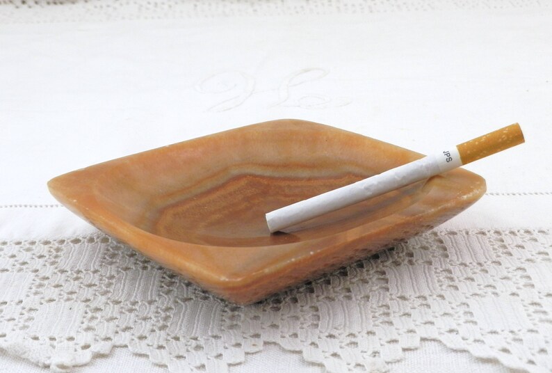 May include: A square, light brown, marble ashtray with a white cigarette resting inside. The ashtray has a slightly raised edge and a smooth, polished surface.