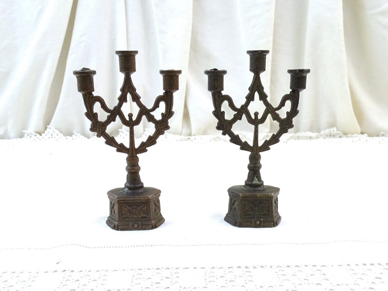 May include: Two antique bronze candle holders with ornate designs. Each holder has three candle arms and a hexagonal base.
