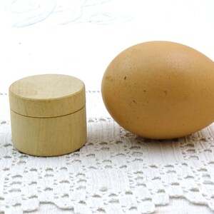 Pode incluir: Uma pequena caixa de madeira com tampa, ao lado de um ovo castanho sobre uma superfície de crochê branca.