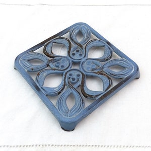 May include: A blue cast iron trivet with a decorative floral design. The trivet is square shaped and has a raised edge.