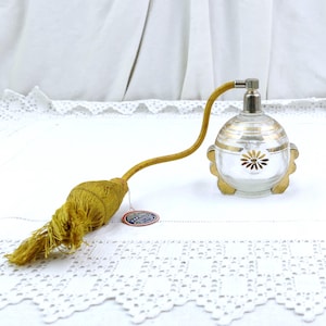 May include: Vintage perfume bottle with a clear glass body, gold accents, and a floral design. The bottle has a long, curved tube and a gold tassel. The bottle is on a white surface.