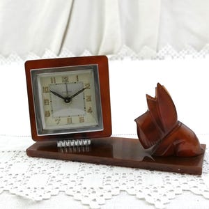 May include: A vintage brown desk clock with a square face and a decorative dog figurine. The clock has a silver face with black hands and numerals. The dog figurine is carved and sits on the same base as the clock.
