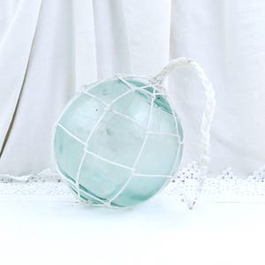 Pode incluir: Uma boia de vidro verde-água, redonda, envolta numa rede branca. Uma corda branca está presa ao topo da boia. Este item decorativo é frequentemente usado em decoração com tema náutico.