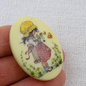 May include: A yellow oval-shaped button with a girl in a yellow hat and a pink and white dress holding flowers. The girl is surrounded by flowers and butterflies.