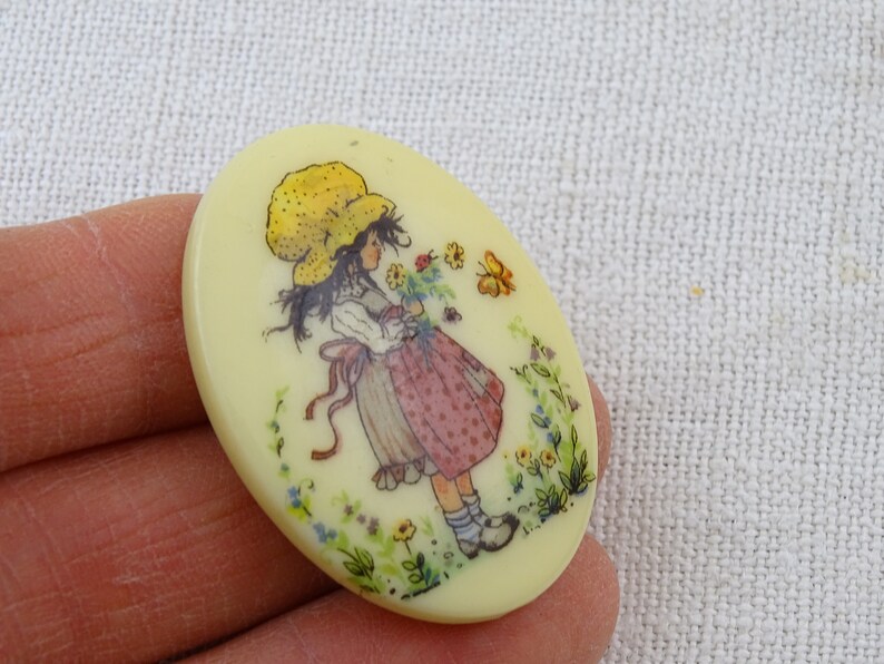 May include: A yellow oval-shaped button with a girl in a yellow hat and a pink and white dress holding flowers. The girl is surrounded by flowers and butterflies.
