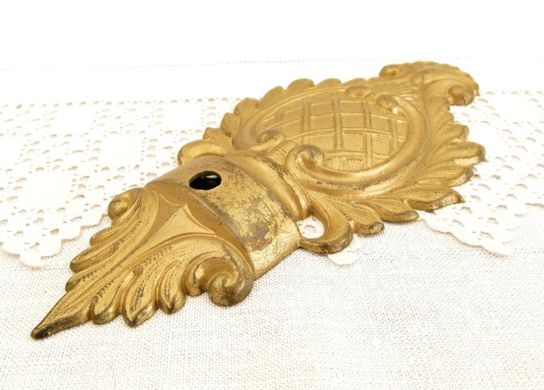 May include: A gold-toned metal decorative ornament with a detailed floral design and a woven pattern in the center. The ornament has a hole in the center, suggesting it may be a wall mount or a part of a larger piece.