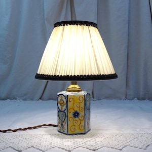 May include: A white lampshade with a black trim sits atop a hexagonal ceramic lamp base. The base is decorated with blue, yellow and green floral patterns.