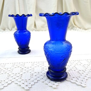 May include: Two cobalt blue glass bud vases with a scalloped rim and an etched floral design. The vases are sitting on a white lace tablecloth.