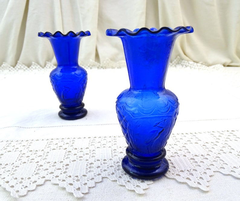 May include: Two cobalt blue glass bud vases with a scalloped rim and an etched floral design. The vases are sitting on a white lace tablecloth.