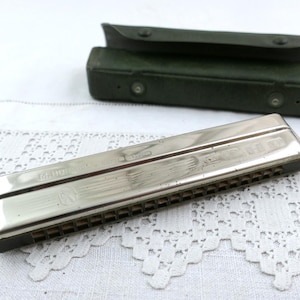 May include: A vintage silver harmonica with a black base and a green case. The harmonica is rectangular with multiple holes and a shiny surface. The case is rectangular with a snap closure. The items are on a white lace tablecloth.